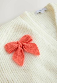 Valge kootud kardigan, millel on vasakul kaeluse lähedal tekstuuriga oranž vibu.