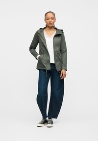 Femme avec des tresses cornrow portant une veste à capuche verte, un haut blanc orné de dentelle, un jean à jambes larges et des baskets noires, debout avec une main dans la poche.