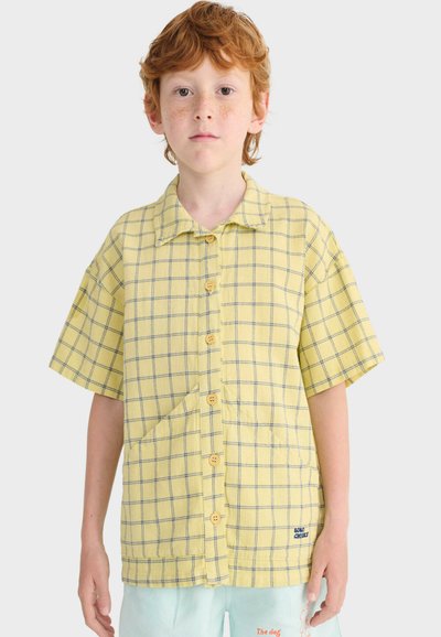 Garçon roux avec des taches de rousseur portant une chemise jaune à manches courtes à boutons avec des lignes bleues en quadrillage et un short bleu clair, sur un fond uni.