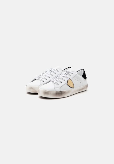 Ζευγάρι λευκά sneakers χαμηλού προφίλ με μαύρα ταμπάκια στη φτέρνα, μπεζ λογότυπο ασπίδας στα πλάγια, λευκά κορδόνια με εκτύπωση και σκόπιμα φθαρμένες σόλες.