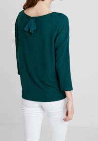 Teal ribbat topp med trekvartslånga ärmar, rund halsringning och en nyckelhålsdetalj bak som är knuten med ett matchande band. Bärs med vita byxor.