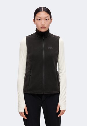 Jeune femme aux cheveux raides porte un gilet noir zippé sur un haut clair à manches longues et un pantalon noir, debout devant un fond uni.