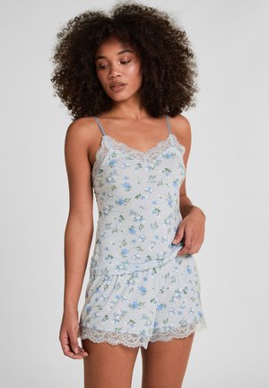 Frau mit lockigen Haaren, die ein hellblaues, mit Spitze verziertes Blumen-Camisole und passende Shorts trägt, vor einem schlichten grauen Hintergrund stehend.