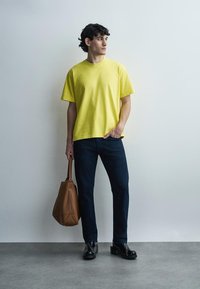 Hombre con camiseta amarilla, vaqueros oscuros, zapatos negros, sosteniendo un bolso de cuero marrón, de pie contra una pared lisa gris claro, mirando hacia la izquierda.