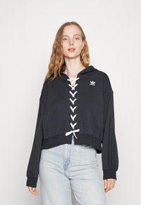 Felpa nera cropped con lacci sulla parte anteriore, occhielli bianchi e cordoncino. Presenta una tasca a marsupio e il logo Adidas sul petto.