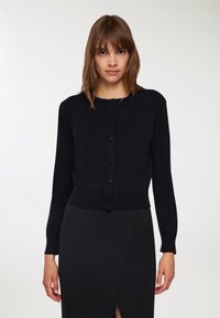 Cardigan lavorato a maglia nero con scollatura rotonda, maniche lunghe e chiusura a bottoni frontale. La texture è liscia con una silhouette aderente.