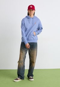 Light blue hoodie with front pocket and adjustable drawstrings, paired with distressed blue jeans featuring a visible patch. Red cap and patterned shoes.