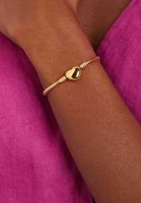 Gouden armband met een gestructureerd ontwerp, met een glanzende hartvormige bedel, gedragen om een pols tegen een roze stoffen achtergrond.