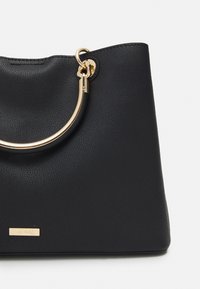 Musta, teksturoidusta nahasta valmistettu käsilaukku, jossa on kultainen pyöreä kahva ja pieni kultainen logolaatta edessä. Muotoilu on jäsennelty ja tilava.