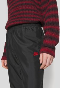 Black nylon pants with an elastic waistband, featuring a red embroidered Nike logo. The worn hand shows a textured red and black striped sweater.