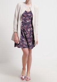 Ljuscreamfärgad långärmad jacka över en mörk blommönstrad klänning med lila, rosa och vita nyanser. Modellen bär glänsande rosa slidesandaler.