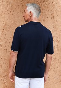 Camisa polo azul marino con cuello acanalado, detalles blancos en los hombros, mangas cortas y un ajuste relajado, combinada con pantalones cortos blancos frente a un fondo texturizado.