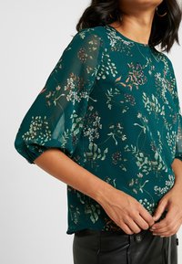 Blouse florale turquoise en tissu transparent, avec des manches bouffantes et un col rond. Le motif comprend des feuilles vertes et de petites fleurs blanches et bordeaux.