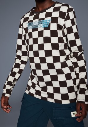 Mężczyzna ubrany w czarno-białą koszulę w kratę z niebiesko-białym logo w paski oraz ciemnoniebieskie spodnie z boczną kieszenią i metką.