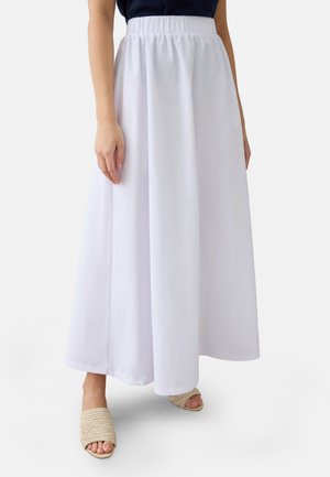 Kvinde iført en lang, hvid nederdel med elastik i taljen og beige åbne flettede slip-ons, stående mod en ensfarvet baggrund.