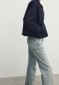 Veste courte bleu marine avec fermeture à boutons, manches oversize, et poches latérales, portée avec un jean droit bleu clair. Texture du tissu lisse.