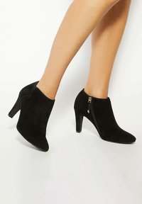 Black suede ankle boots with pointed toes, 9 cm heels, and side zippers featuring gold accents. Smooth texture with a sleek design.
