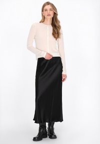 Lange zwarte satijnen rok gecombineerd met een lichtbeige blouse met lange mouwen en knopen. Het model draagt zwarte enkellaarsjes en staat tegen een witte achtergrond.