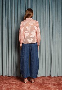 Žena s dlhými vlasmi stojí čelom k modrým závesom, oblečená v priehľadnej ružovej kvetinovej blúzke, širokých námorníckych nohaviciach so trblietkami a bielych vysokých opätkoch.