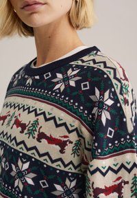 Pletený svetr s vánočním vzorem v červených a zelených akcentu, sněhovými vločkami a jezevčíky na smetanovém a námořnickém pozadí.