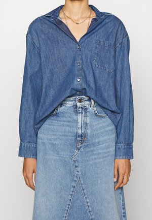 Kvinde iført en løs blå denimskjorte, der er stoppet ned i en højtaljet lys blå denimnederdel med frontslids og knaplukning.