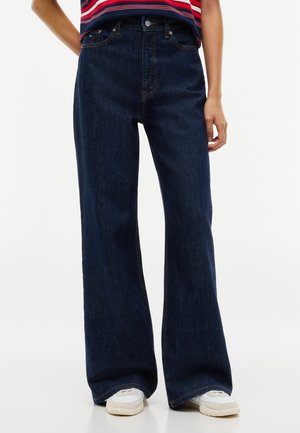 Personne portant un jean bleu foncé à jambes larges et des baskets blanches, debout avec une main près de la hanche.