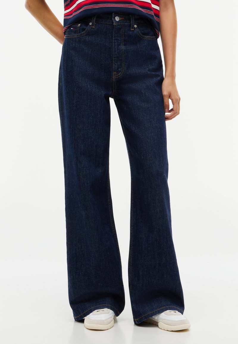 Personne portant un jean bleu foncé à jambes larges et des baskets blanches, debout avec une main près de la hanche.