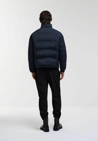 Jachetă puffer de marină cu cusături orizontale, guler înalt, manșete elastice și o croială lejeră, asortată cu pantaloni negri și cizme.