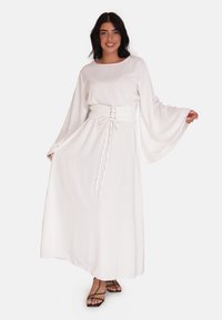 Robe longue blanche avec des manches cloche fluides, une taille cintrée et des détails lacés. Tissu lisse avec une coupe décontractée, associée à des sandales noires à lanières.