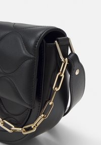 Borsa a mano in pelle nera con motivo a onde trapuntate, tracolla a catena color oro, chiusura con patta magnetica e texture liscia.