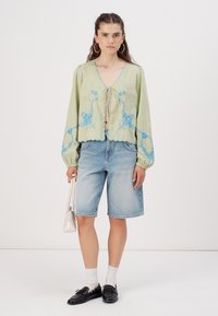 Ljusgrön blus med blå blommönsterbroderi, v-ringad och med knytning framtill, ihop med denimshorts. Tillbehör inkluderar en vit handväska och svarta loafers.