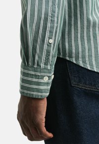 Groen gestreept shirt met witte lijnen, knoopmanchetten en geborduurd logo. De stof lijkt zacht met een casual pasvorm, gecombineerd met donkere denim.