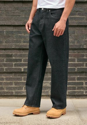 Homme portant un jean noir ample, des bottines de travail marron clair et une chemise blanche, debout contre un mur en briques grises sur une surface pavée.