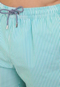 Pantaloni corti a righe azzurro chiaro e bianchi con vita elastica e dettaglio di cordoncino, caratterizzati da una cravatta decorata e punte metalliche.