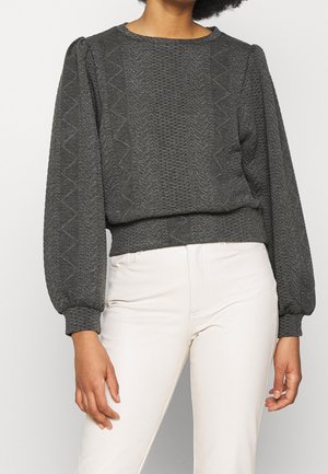 Vrouw draagt een donkergrijze gebreide trui met textuur en ballonmouwen, gecombineerd met een witte broek, tegen een effen achtergrond.