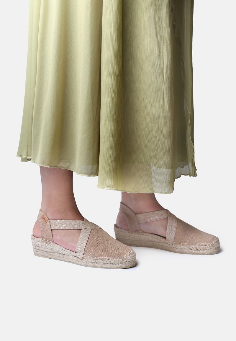 Voeten met beige espadrille sleehak sandaal met gekruiste bandjes, gecombineerd met een lichtgroene geplooide rok tegen een effen achtergrond.