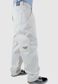 Pantaloni cargo bianchi in denim con ampie tasche laterali, logo ricamato e vestibilità rilassata, abbinati a sneaker nere e blu.
