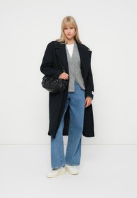 Largo abrigo negro, chaleco gris, camiseta blanca, jeans de pierna ancha azules y zapatillas blancas. Sostenido un bolso negro.