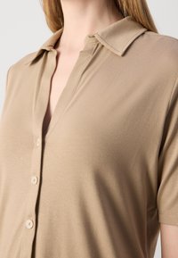 Polo beige à manches courtes avec un col en V, fermeture à boutons et une texture douce et lisse. Le col est légèrement structuré.