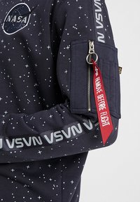 Felpa navy con stampa a stelle, dotata di tasca con zip sul maniche, testo grigio e un'etichetta rossa rimovibile con la dicitura "RIMUOVERE PRIMA DEL Volo."