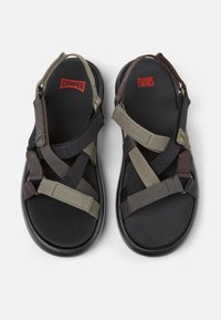 Utomhussandaler med svart sula. Ovandelen har grå och bruna tygremmar som korsar en svart fotbädd, märkt med "CAMPER" och "TWINS."