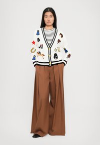 Donna che indossa un cardigan bianco con toppe a forma di cavallo e nastro, una camicia a righe, pantaloni larghi marroni e scarpe nere, in piedi contro uno sfondo bianco.