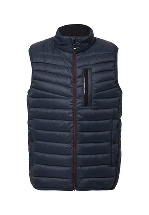 Tumši zila polsterētā vestē ar augstu apkakli, priekšējo rāvējslēdzēju un rāvējslēdzēja krūšu kabatu. Iezīmējas ar kontrastējošo sarkano rāvējslēdzēju un plakano šuvēm.