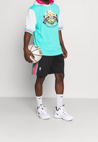 Lichtblauw mesh hoodie shirt met witte mouwen, zwarte shorts met kleurrijke zijpanelen en witte sneakers, die een basketbal vasthoudt.