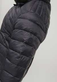 Giacca a vento nera, con un design trapuntato e tessuto testurizzato. Include un logo circolare sulla manica per la personalizzazione.