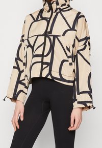 Giacca a vento beige tagliata con motivo astratto nero, chiusura con zip, colletto alto e polsini elasticizzati. Indossata sopra leggings neri attillati.