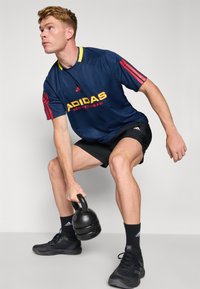 Tamsiai mėlyna Adidas sportinė marškinė su raudonomis juostelėmis, geltonu apykakle ir logotipu. Modelis dėvi juodas šortus ir batus, laikydamas juodą ketlebell'ą.