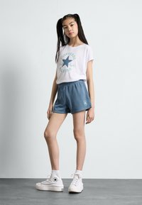 Bílá krátká tričko s krátkým rukávem a tyrkysovým potiskem "Converse All Star", spárované s modrými šortkami a bílými vysokými teniskami. Hladká bavlněná tkanina.