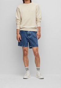 Béžová mikina s malým logem, kombinovaná s modrými denimovými šortkami a bílými teniskami. Ponožky mají černý nápis. Jednoduchý, ležérní outfit.