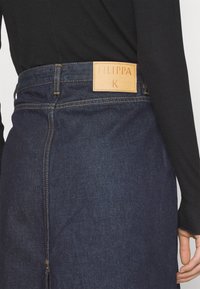 Jupe en jean bleu foncé avec des coutures jaunes et un patch en cuir beige marqué "FILIPPA K", portée avec un haut noir à manches longues.